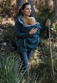 Türkisfarbene Softshelljacke mit grauen Akzenten, mit Reißverschluss versehenen Taschen, über einem gelben Strickoberteil getragen. Eine Frau hält ein Kind in einem Tragehilfe vorne und einen Gehstock.