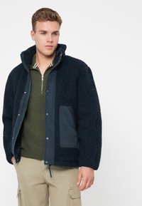 Mladic nosi tamnoplavu flis jaknu preko maslinasto zelene majice s patentnim zatvaračem i bež cargo hlačama, stoji naslonjen na bijelu pozadinu.