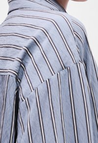 Gros plan sur une personne portant une chemise bleu clair avec des rayures blanches verticales et marron foncé, montrant les détails des épaules et du cou.