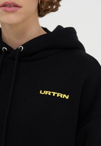 Černá mikina s kapucí se žlutým logem "URTRN" na přední straně. Má šňůrky na stažení, měkkou texturu a pohodlný volný střih.