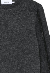 Mørkegrå striktrøje med en tekstureret strik, der har sort frynse-detalje langs ærmet. Klassisk rund halsudskæring, afslappet design.