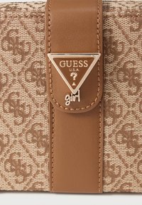 Hnedá textilná taška s vzorovanou tkaninou a koženým remienkom. Obsahuje trojuholníkové kovové logo s nápisom "GUESS" a doplnkom "girl".
