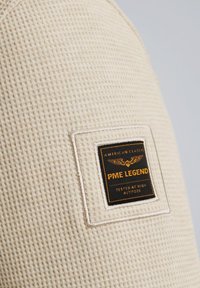 Beżowa tkanina o fakturze z wzorem wafelka, z czarną i złotą prostokątną metką z napisem "PME LEGEND" i wzorem orła.