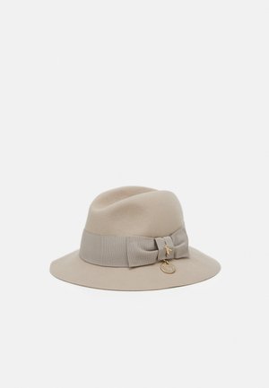 Beige fedora-hoed met brede rand, bijpassende stoffen band, sierlijke strik en twee kleine goudkleurige bedeltjes aan de strik bevestigd.