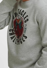 Harmaa collegepaita, jossa suuri graafinen hahmoprintti polkupyörällä, ympärillä teksti "Obstacles Challenges". Klassinen pyöreä kaula-aukko.