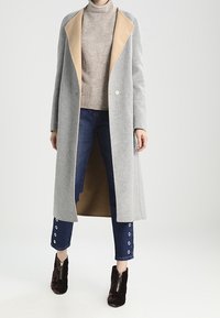 Person, der einen langen grauen Mantel mit beigem Futter, einen beigen Rollkragenpullover, cropped blaue Jeans mit Ösen und dunkelbraune Stiefeletten mit Frontreißverschluss trägt.