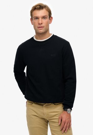 Sort sweatshirt med ribbede manchetter og rund halsudskæring, præget med et lille logo på brystet. Sammen med lyse khaki bukser og et ur.