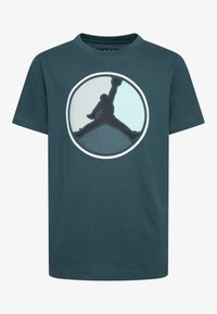 Teal cotton t-paita, jossa on pyöreä logo, jossa on musta siluetti hyppäävästä hahmosta ja vaaleansinisiä yksityiskohtia. Lyhyet hihat ja pyöreä kaula-aukko.