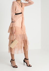 Kvinna klädd i en persikofärgad klänning i spets med lager och svart kant, håller en beige kuvertväska och bär svarta högklackade sandaler, stående mot en vit bakgrund.