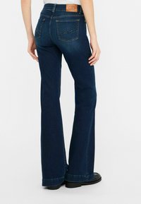 Jeans de campana azul oscuro hechos de denim, con un parche de cuero marrón en la cintura, bolsillos cosidos y un dobladillo limpio.