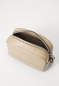Borsa in pelle beige con logo "Calvin Klein Jeans" in rilievo, chiusura con zip e una texture liscia. L'apertura superiore mostra la fodera interna.
