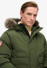 Grøn polstret parka-jakke med en hætte foret med pels, lynlås foran og to forlommer. Har et logo-patch på venstre ærme.