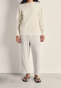 Krämfärgad fleece-tröja med ribbade ärmar, tillsammans med ljusgrå loungbyxor och blå flip-flops, som ger en avslappnad passform och mjuka texturer.