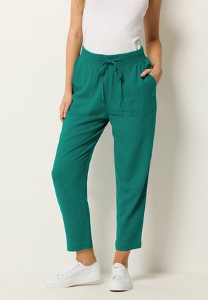 Vrouw draagt teal taps toelopende broek met koordsluiting in de taille en voorzakken, gecombineerd met witte sneakers en een witte mouwloze top.