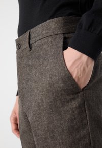Pantaloni marroni con texture, vita aderente, bottoni e tasche laterali; realizzati in tessuto tessuto, che mostrano un primo piano del materiale e del design.