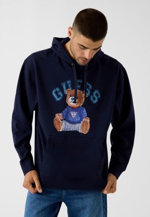 Man draagt een donkerblauwe hoodie met een teddybeerafbeelding en de tekst "GUESS", gecombineerd met blauwe spijkerbroek, kijkt naar beneden tegen een effen achtergrond.