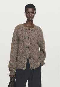 Cardigan lavorato a maglia marrone con texture, scollo rotondo, chiusura a bottoni e maniche lunghe. Indossato su una maglietta scura, abbinato a pantaloni larghi.