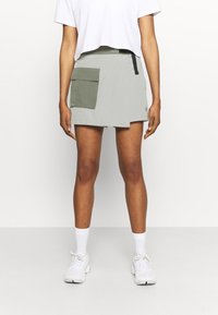 Falda verde claro con un diseño de envoltura, que cuenta con un gran bolsillo frontal. Hecha de material ligero, combinada con una camiseta corta blanca y zapatillas blancas.