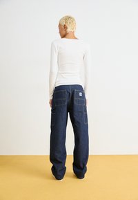 Dark blue denim wide-leg pants with multiple pockets and yellow stitching, paired with a fitted long-sleeve white ribbed top.