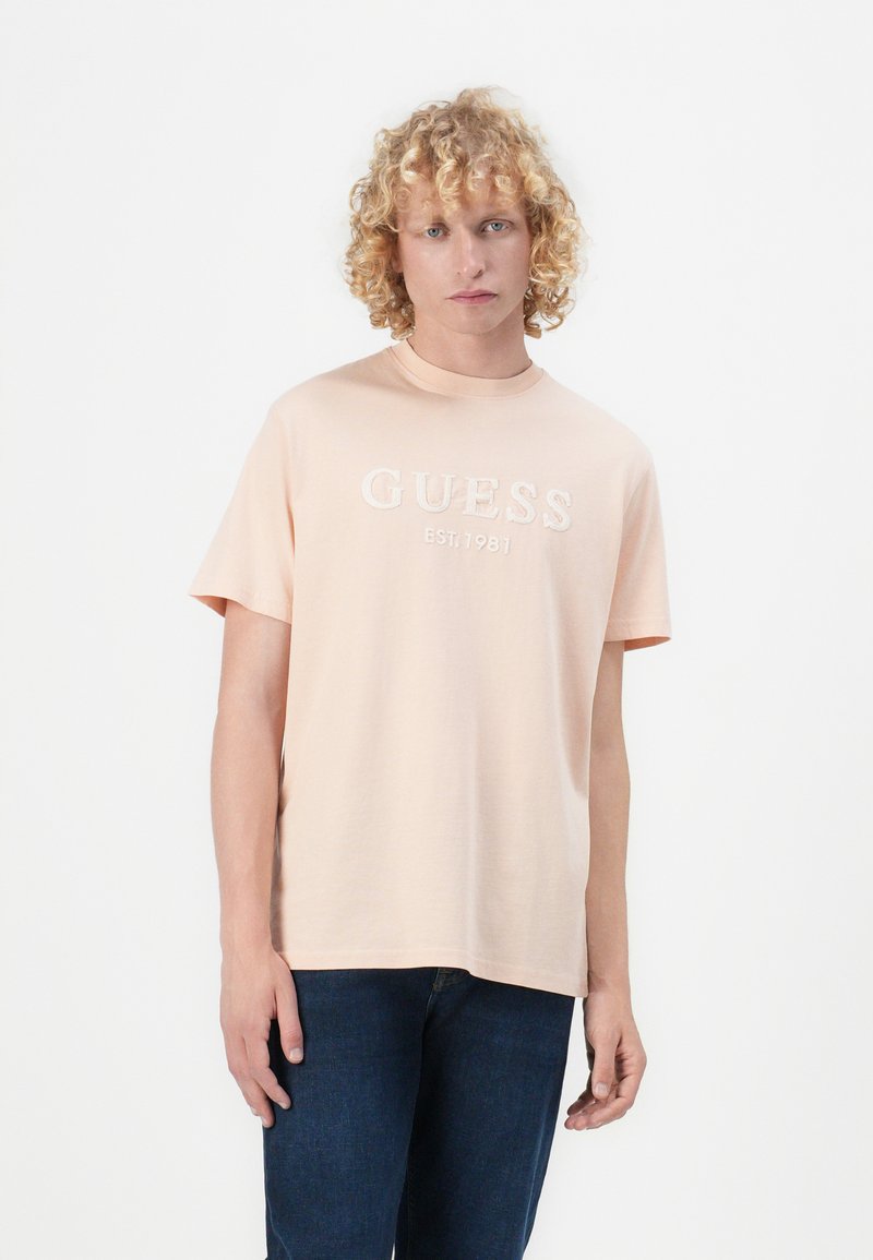 Svijetloružičasta pamučna majica s kratkim rukavima, s teksturiranim logom "GUESS" i "EST. 1981" u bijeloj boji, okrugli izrez, klasičan kroj.