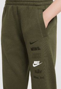 Olive green sweatpants featuring various Nike logos in black and white, side pocket, elastic waistband, and a soft texture.