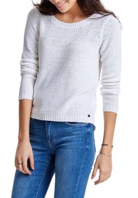 Pull en tricot blanc avec un col rond et des manches longues. La texture est en maille lâche et un petit logo noir est visible au niveau de l'ourlet.