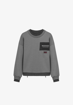 Sweatshirt gris avec une poche à rabat noire, col et poignets côtelés, arborant le texte "RÉSONANCE" et une étiquette en bas. Tissu texturé.