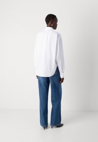 White long-sleeve shirt, relaxed fit with back pleat, paired with wide-leg blue denim jeans, and black pointed heels. Simple design, no patterns.