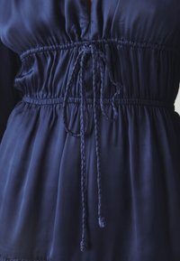 Námořnické modré šaty s nařaseným pasem, které mají nastavitelné červené tkaničky a hladkou, hedvábnou texturu.
