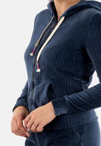 Sudadera con cremallera de color azul marino, de canalé, con capucha, cordones color crema con acentos en rosa y bolsillos frontales. Material suave y texturizado.