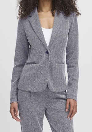 Woman wearing a gray herringbone patterned suit jacket and matching pants over a white top, standing with arms relaxed by her sides.