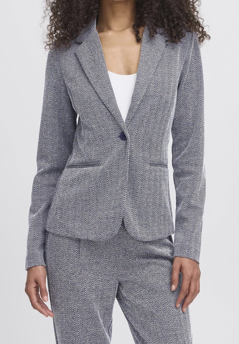 Femme portant une veste de costume grise à motif chevrons et un pantalon assorti par-dessus un top blanc, debout, les bras détendus le long du corps.