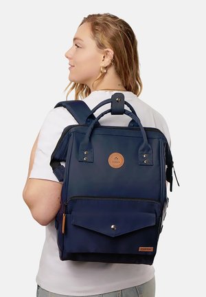 Frau mit mittellangen Haaren, die ein weißes Hemd trägt und einen dunkelblauen Rucksack mit Fronttasche und oberen Griffen trägt.