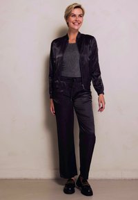 Schwarze Satin-Bomberjacke über einem grauen, strukturierten Oberteil, kombiniert mit schwarzen, weit geschnittenen Hosen. Massive schwarze Schuhe mit goldenen Akzenten vervollständigen das Outfit.