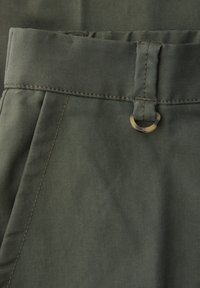 Nærbillede af olivengrønne bukser med linning, bæltestrop og lille rundt skildpaddeskalringdetalje på stoffet.