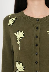 Oliivi vihreä neuletakki mustilla napilla, jossa on keltaista kukkaihastusta edessä. Ribbireunus kauluksessa ja pehmeä neulepinta.