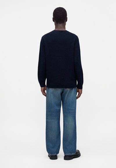 Personne debout de dos portant un pull bleu marine foncé, un jean bleu ample et des chaussures noires sur un fond clair uni.