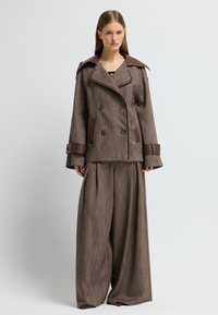 Giacca doppiopetto marrone con texture, colletto ampio e dettagli in pelle marrone, abbinata a pantaloni larghi coordinati dello stesso colore.