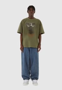Oliven grøn oversized t-shirt med et grafisk design af en engel og teksten "Guardian Angel"; parret med løstsiddende blå denim jeans.