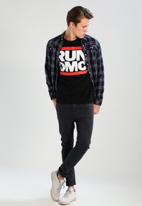 Neformální outfit skládající se z černého trička s odvážným bílým a červeným grafickým textem, přes které je navlečená modrá kostkovaná košile, a tmavých džínů. Bílé tenisky.