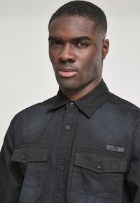 Svart skjorte med knapper laget av bomull, med to brystlommer med klaffer, spiss krage, og en subtil glans på stoffet.