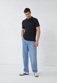 Polo noir à manches courtes, jean droit bleu clair et baskets noires et blanches. Le mannequin se tient devant un fond neutre.