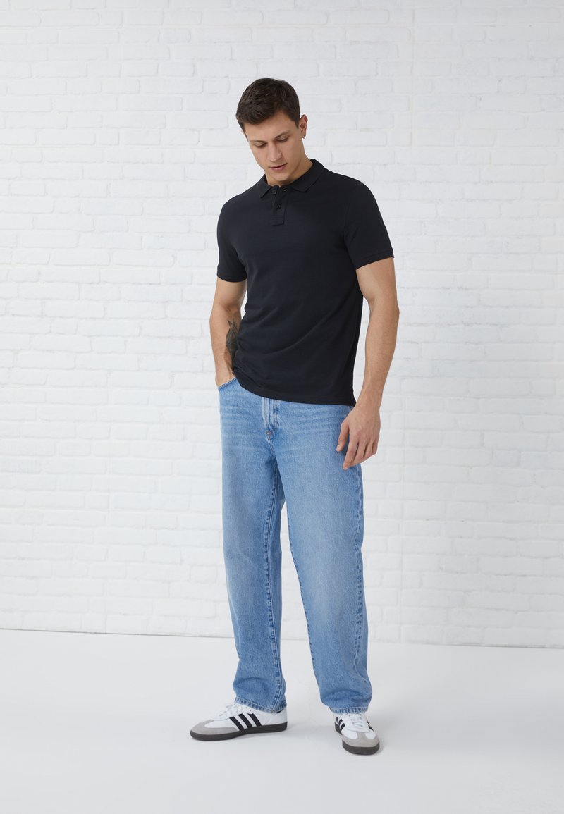 Polo noir à manches courtes, jean droit bleu clair et baskets noires et blanches. Le mannequin se tient devant un fond neutre.