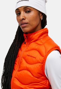 Oranje gewatteerde vest met een hoge kraag, voorzien van het merk "Peak Performance" in het rood. Draag het over een witte lange mouwen top, met een ritssluiting.