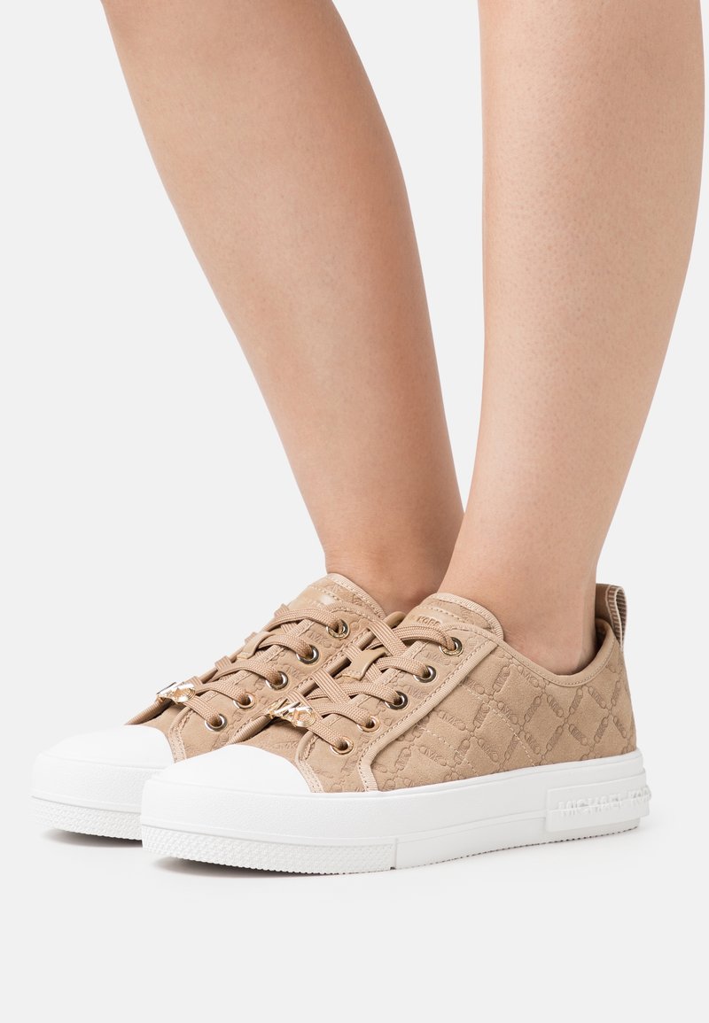 Pieds portant des baskets basses beiges avec semelles blanches et motif matelassé, visibles sur un fond blanc uni.