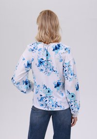 Blusa bianca con motivo floreale blu, con scollatura a balze, maniche lunghe e polsini elasticizzati. Indossata con jeans blu scuro.