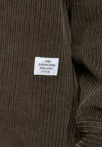 Rudų grūdėtos medžiagos audinys su vertikaliomis raukšlėmis, kuriame matomas baltas etiketė su užrašu "J. Jones Superior Goods Denim Supply Clth. Co."
