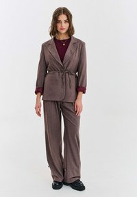 Hnedé priliehavé sako s opaskom a širokými nohavicami. Má zapínanie saka na gombíky a manžety s kontrastnými burgundskými detailmi.