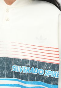 Bílá polo tričko s třemi knoflíky, s vodorovnými pruhy v červené, modré a tmavě modré barvě. Na spodní části je v tučném modrém písmu nápis "SILVERADO SPIKE".