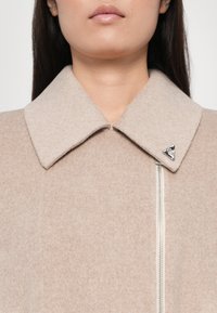 Beige wollen jas met grote kraag, zilveren rits en driehoekig logo accent. Zachte textuur, schoon ontwerp met minimale detailing.